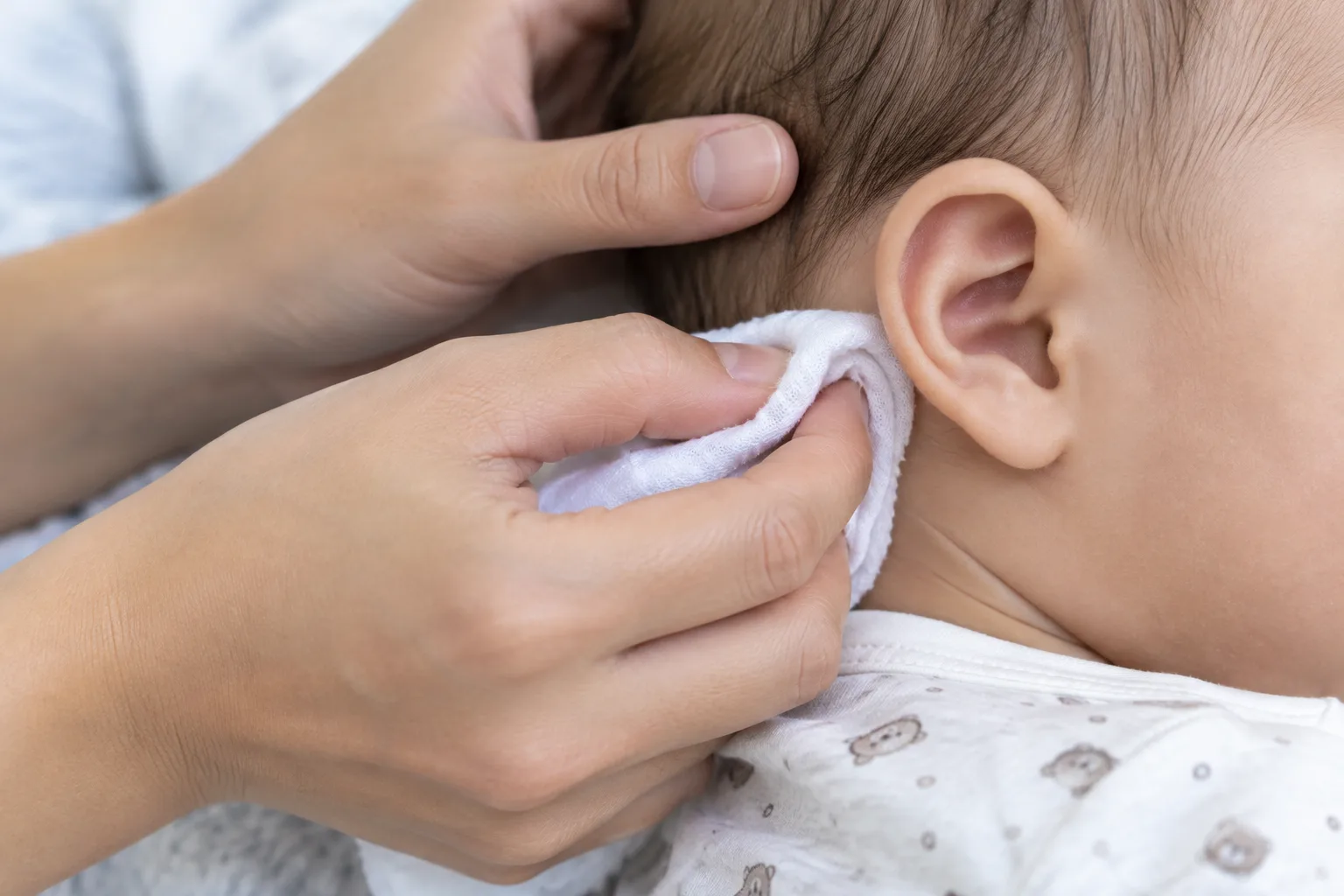 Un parent passe délicatement un linge humide sur le pavillon et les plis visibles derrière l’oreille de bébé.