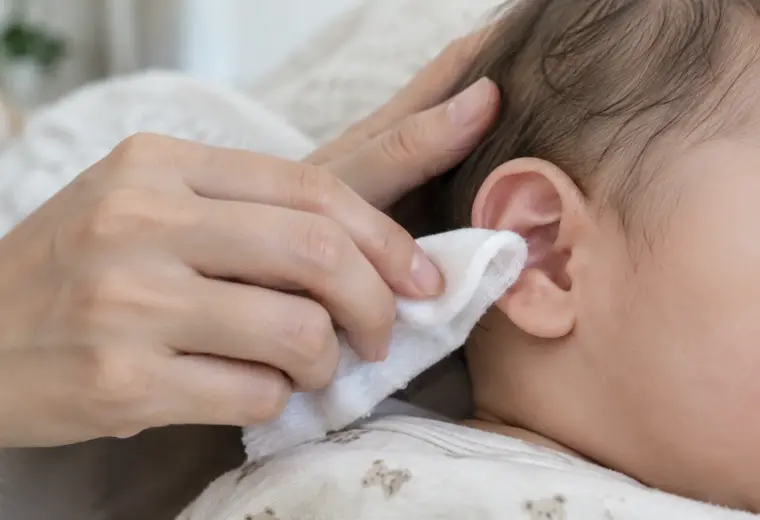 Une main nettoie doucement le contour de l’oreille d’un bébé avec un linge humide.